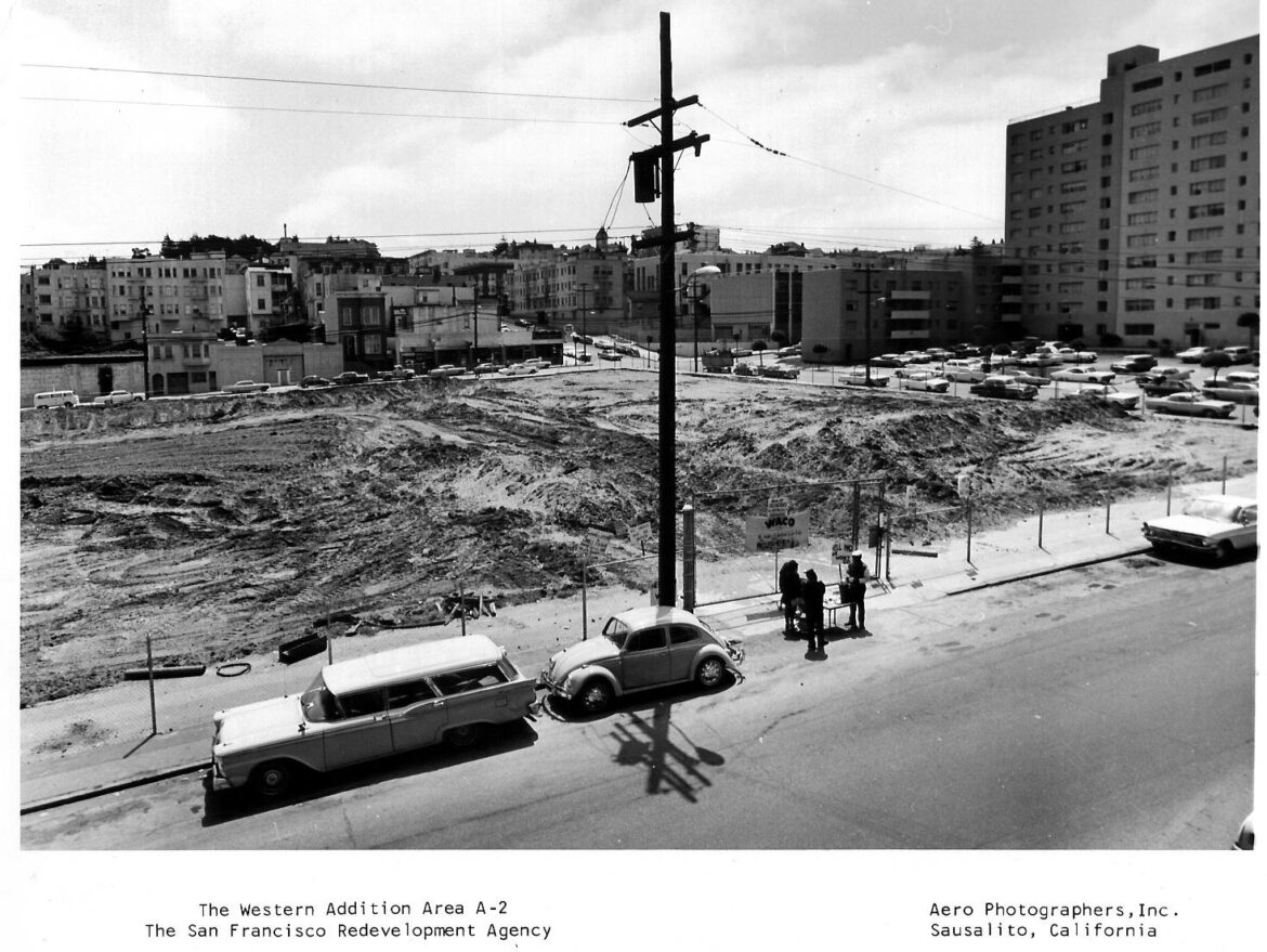 An aerial black and white photo displays an empty dirty lot surrounded by chain link fences. Two cars are parked next to the sidewalk in front of the lot. Behind the cars, a group of three people stand around a small table.