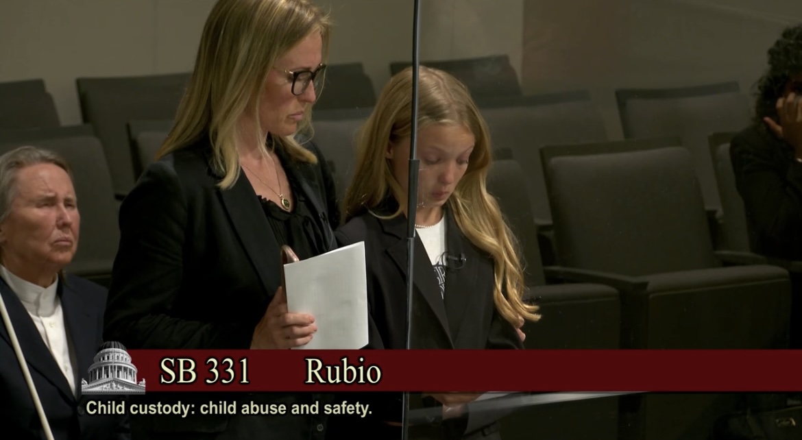 A girl dressed in black stands at a lectern where she is speaking. Her mother, also dressed in black, stands beside her. A few people sit behind them in theater-style seats.