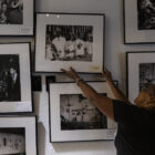 A woman with a long black ponytail reaches up to straighten the frame of one of many black and white photographs displayed in a closely spaced array on a wall in an art gallery.