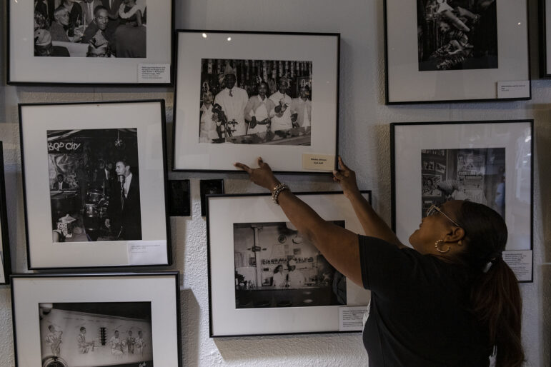 A woman with a long black ponytail reaches up to straighten the frame of one of many black and white photographs displayed in a closely spaced array on a wall in an art gallery.