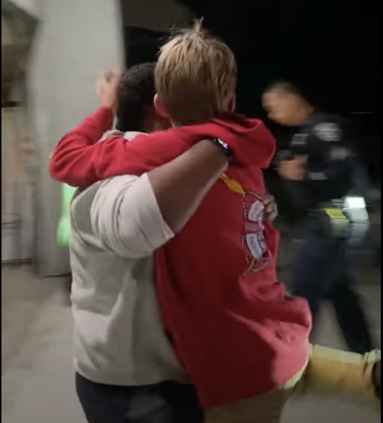 An adult forcibly picks up a boy wearing a red sweatshirt with a hood. Their faces are turned away. A police officer in uniform walks in the background.