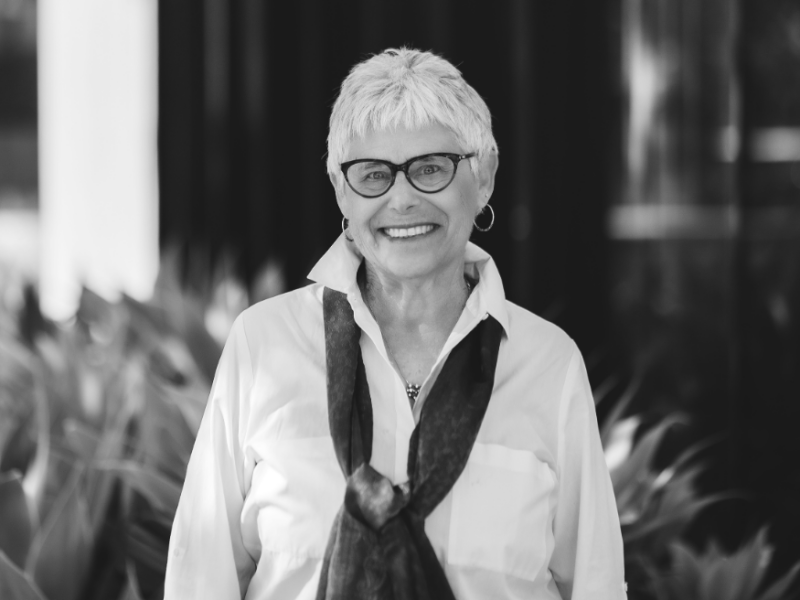 Black and white image of a woman with short silver hair wearing dark-framed eyeglasses, a light colored blouse and a dark scarf standing in front of leafy foliage. The background is slightly blurred.
