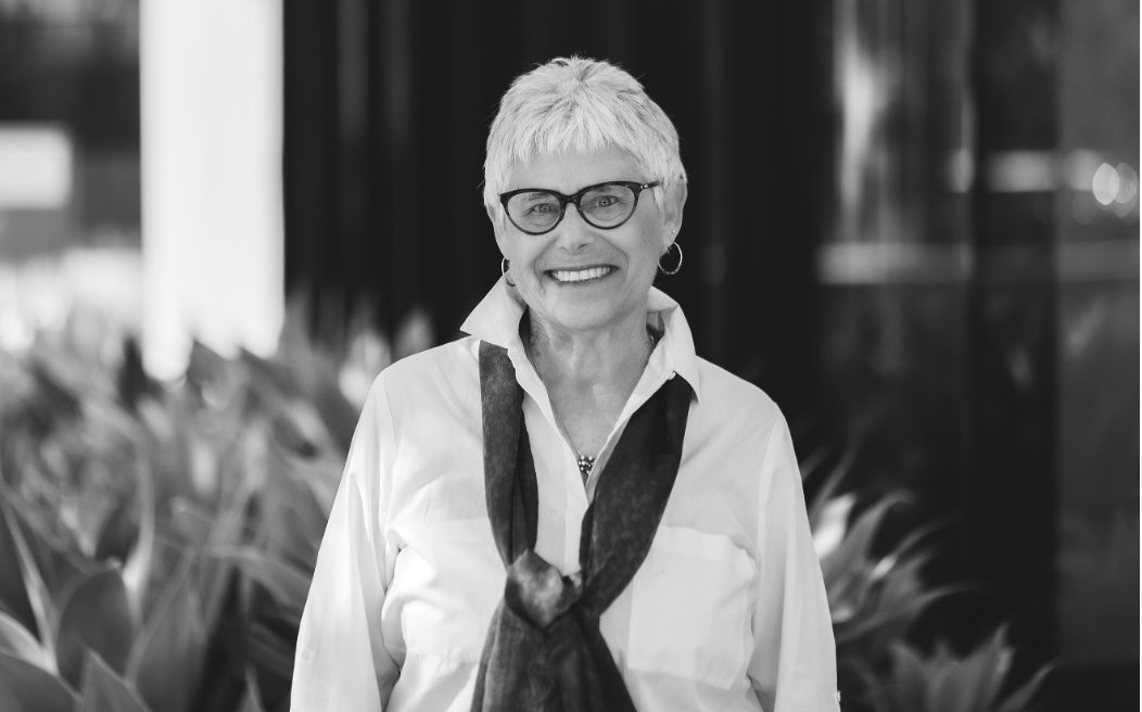 Black and white image of a woman with short silver hair wearing dark-framed eyeglasses, a light colored blouse and a dark scarf standing in front of leafy foliage. The background is slightly blurred.