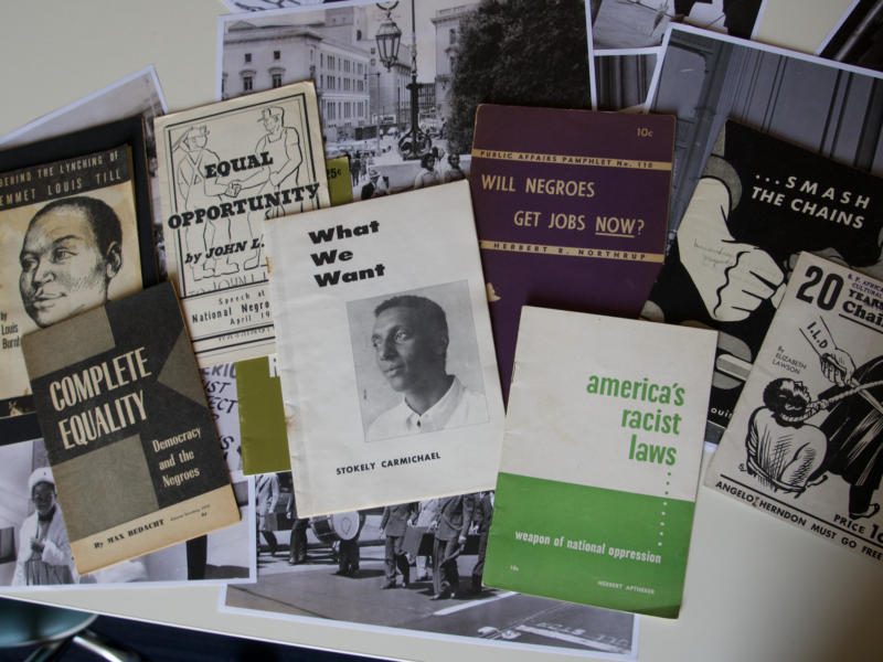 Eight historical booklets are spread out on top of several black and white photos of Black Americans in San Francisco protesting for civil rights in the 1960s. The pamphlet at the center of the table is titled "What We Want" and has a black-and-white photo of it's author Stokely Carmichael, who later changed his name to Kwame Ture. To its right is a lime green and white pamphlet called "America's Racist Laws" by Herbert Aptheker. Other pamphlet titles include "Behind the Lynching of Emmet Louis Till," ""Complete Equality: Democracy and the Negroes," and "Will the Negro Get Jobs Now?".