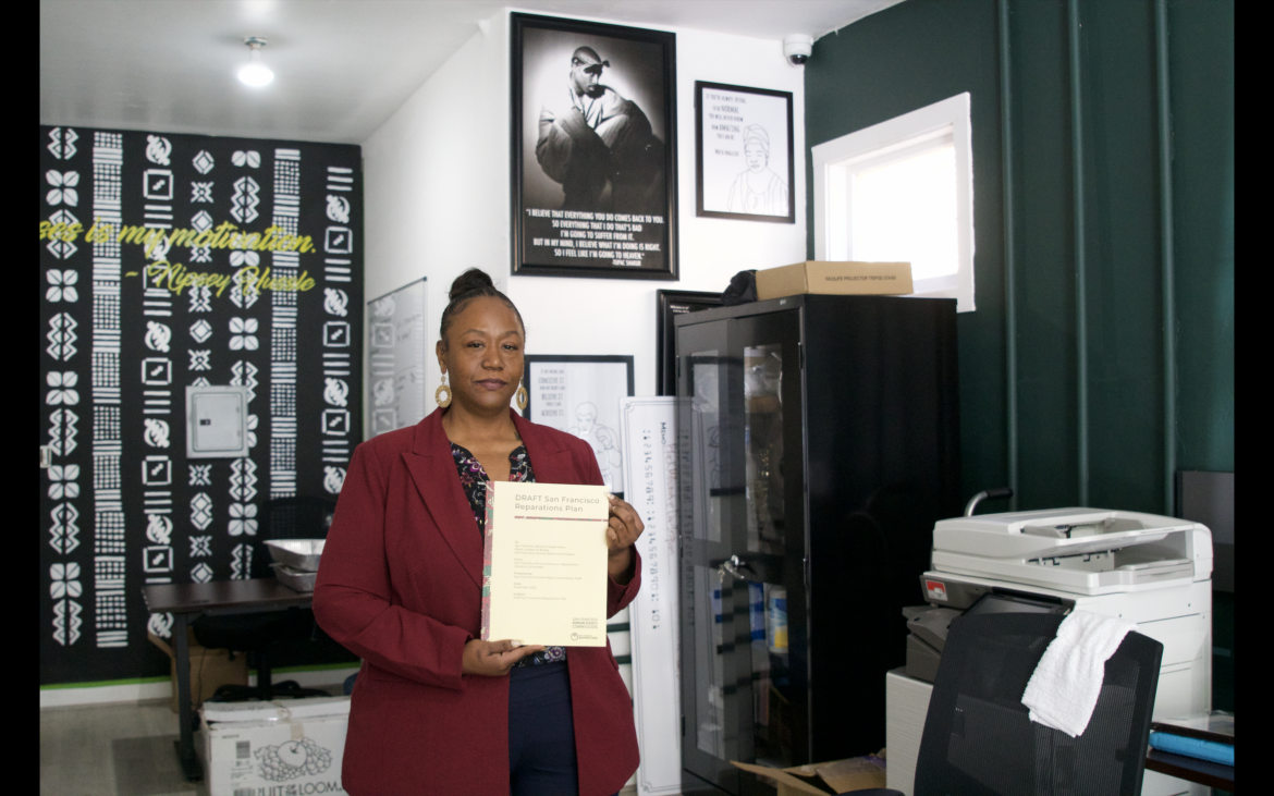 An African American woman in a burgundy jacket and blue pants is standing in an office space with several posters on the wall behind her. She holds a cream-colored booklet with the words "DRAFT San Francisco Reparations Plan" written across the top.