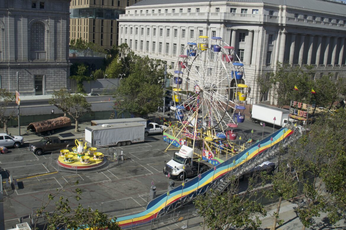 Three colorful carnival attractions — a Ferris wheel, a wavy slide and a tea cup ride — are being set up on a street in San Francisco's Civic Center neighborhood. Several trucks are parked nearby.