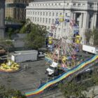 Three colorful carnival attractions — a Ferris wheel, a wavy slide and a tea cup ride — are being set up on a street in San Francisco's Civic Center neighborhood. Several trucks are parked nearby.