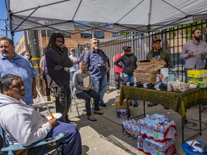 People stand and sit under and around a white shade structure. There is a table stacked with pizza boxes and other snacks, with three large bundles of bottled water underneath.