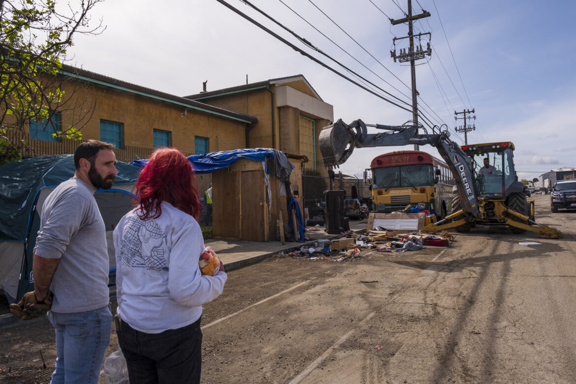 A man and a woman stand in the street talking with their backs toward the camera. In the background, a backhoe operator prepares to use his machine to pick up trash and discarded items that have been pushed into the street in front of an old yellow school bus.