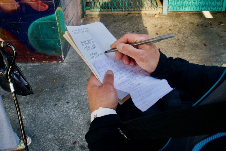 A close-up shows a man's hands as he copies a list of phone numbers and addresses onto a piece of paper.