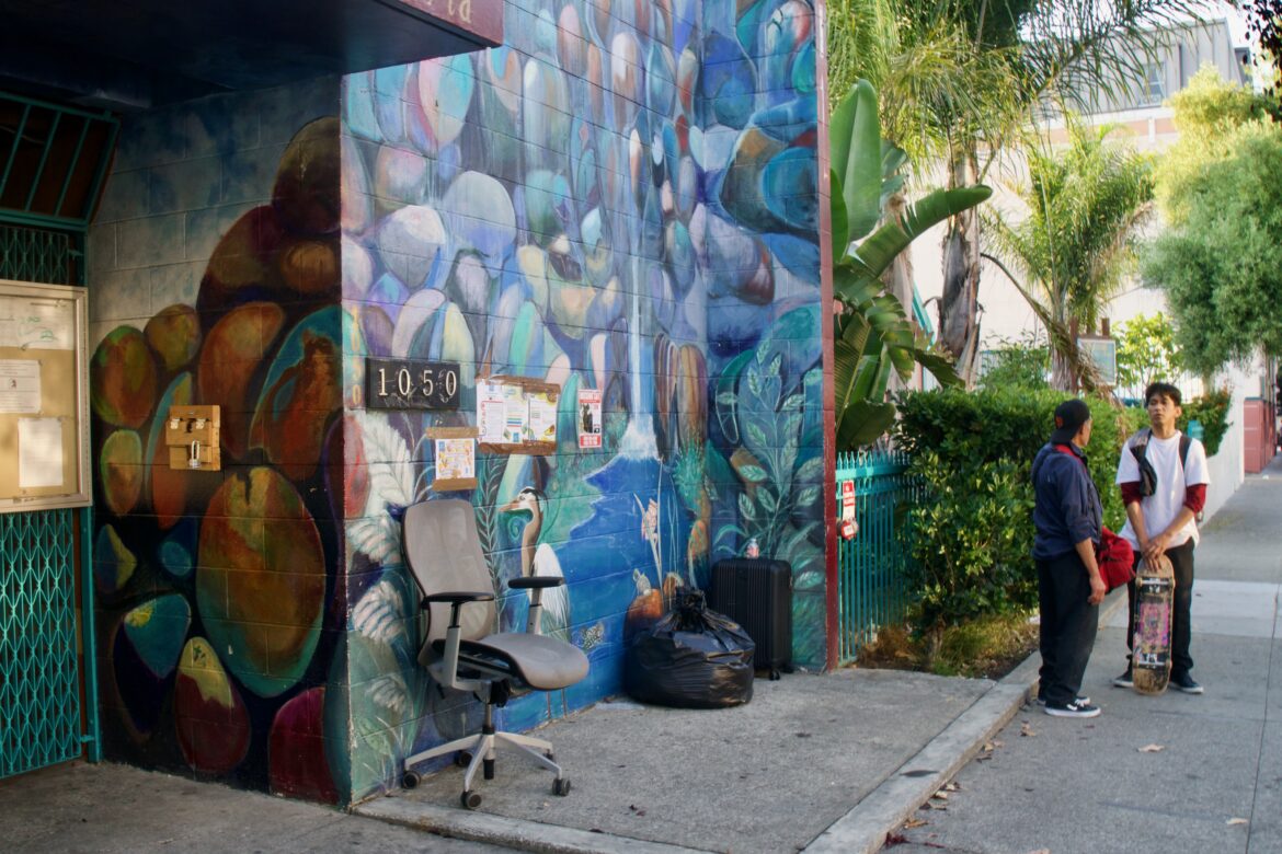 A man in a white shirt holding a skateboard speaks to another man on the sidewalk. In front of a colorful building nearby, a grey office chair rests next to a black trash bag and suitcase.