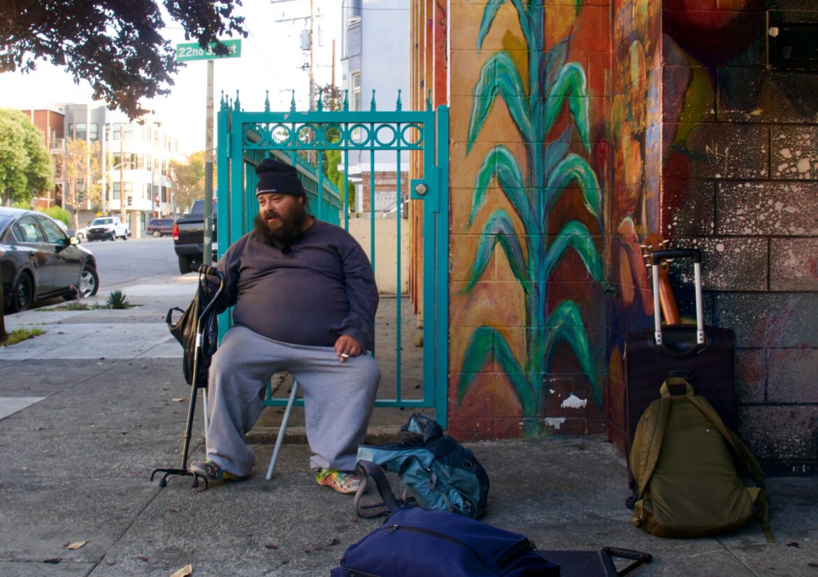 A man in gray sweatpants, a long sleeved shirt and a black beanie sits on a chair with his hand on his cane. To his left, several backpacks and suitcases are scattered on the sidewalk.