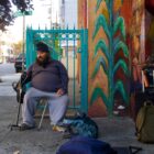 A man in gray sweatpants, a long sleeved shirt and a black beanie sits on a chair with his hand on his cane. To his left, several backpacks and suitcases are scattered on the sidewalk.