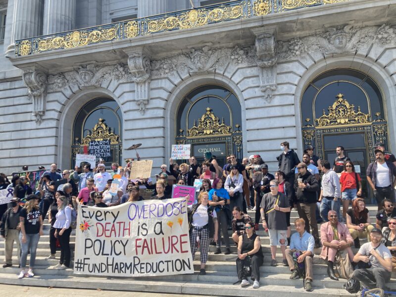 A rally at City Hall with a sign reading "Every Overdose Death Is a Policy Failure" #fundharmreduction