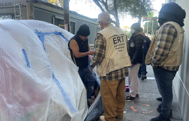 Three people wearing vests and jackets with city logos speak with a man standing in the entrance of a tent covered with plastic tarps on a city sidewalk.