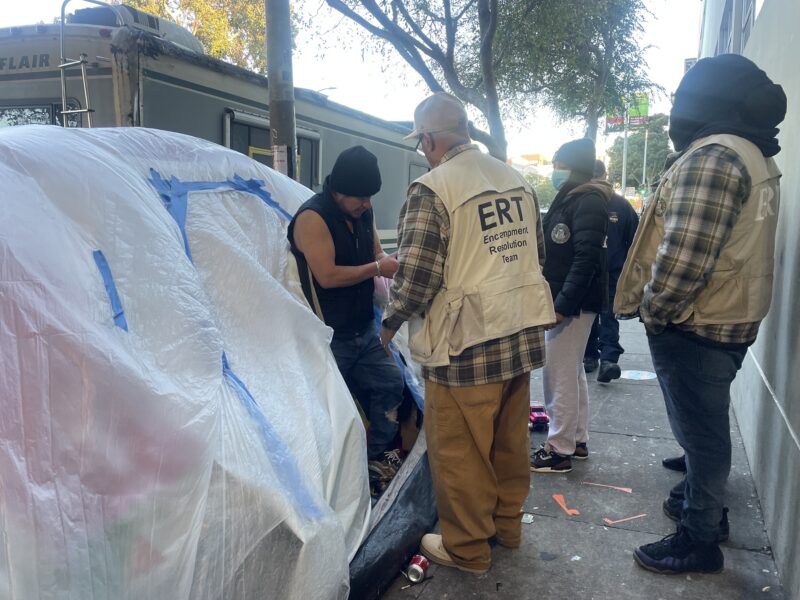 Three people wearing vests and jackets with city logos speak with a man standing in the entrance of a tent covered with plastic tarps on a city sidewalk.