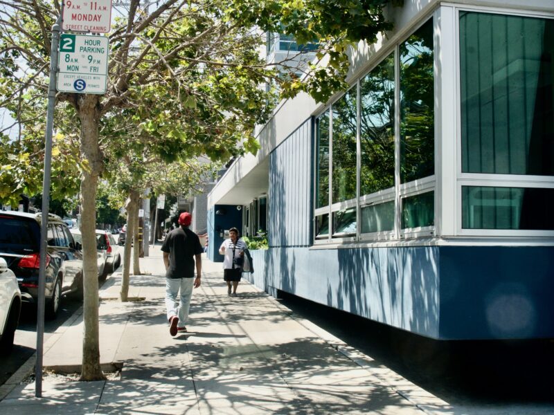 A man with a red hat and black shirt with his back towards the camera walks down the sidewalk. To his right, a woman in a white shirt strolls past a blue and white building with large glass windows.