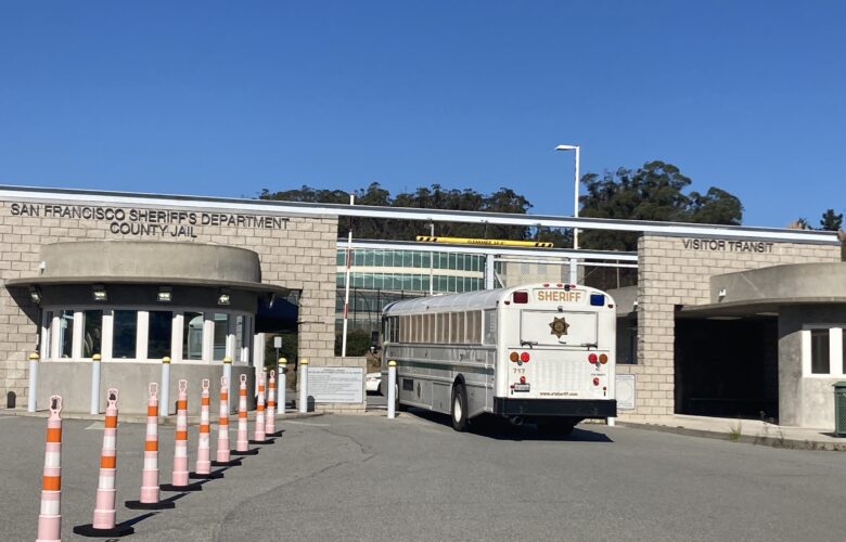 A San Francisco Sheriff's Department prisoner transport bus arrives at the San Francisco County Jail in San Bruno. The cream-colored bus is driving through the entrance, which is flanked by two brown brick walls. A series of tall, narrow traffic cones appear on the left side of the frame. District Attorney Brooke Jenkins criticizes the use of pretrial diversion programs offering defendants accused of selling drugs rehabilitation, counseling and training rather than jail sentences. Many such suspects are held at the San Francisco County Jail in San Bruno.