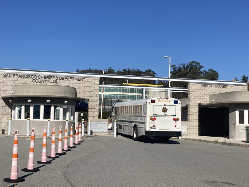 A San Francisco Sheriff's Department prisoner transport bus arrives at the San Francisco County Jail in San Bruno. The cream-colored bus is driving through the entrance, which is flanked by two brown brick walls. A series of tall, narrow traffic cones appear on the left side of the frame. District Attorney Brooke Jenkins criticizes the use of pretrial diversion programs offering defendants accused of selling drugs rehabilitation, counseling and training rather than jail sentences. Many such suspects are held at the San Francisco County Jail in San Bruno.