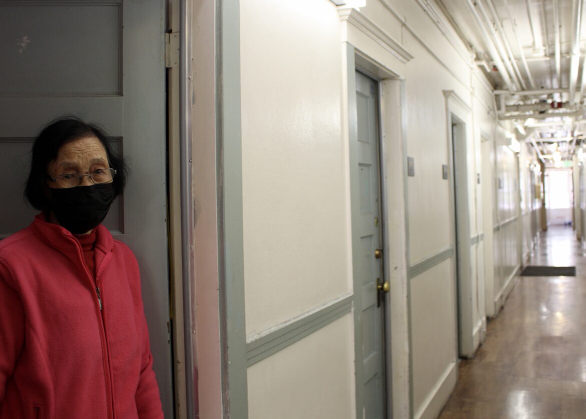 A woman in her 80s wearing a pink fleece jacket and a black face mask stands in her doorway in a single-room occupancy building in Chinatown.