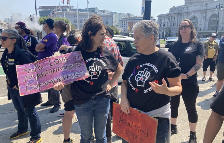 Two women are speaking to each other at a protest rally on a plaza outside of San Francisco City Hall. They are Supervisor Hillery Ronen, who holds a pink sign that reads "We Love Marginalized Communities," and Vitka Eisen, CEO of HealthRight 360, who holds a sign that reads "Harm Reduction Works." They are both wearing jeans and black T-shirts. Many of the people standing around them are similarly dressed.