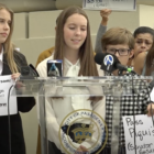At a news conference about the passage of Piqui's Law, three teenage girls and a middle school-aged boy stand behind a clear lectern holding microphones from news outlets and bearing the seal of the city of Pasadena. They and others in the crowd hold signs, some of which read "Governor Newsom: Please sign SB 331 (Rubio) Piqui's Law" and "Pass Piqui's Law (Senator Susan Rubio)." They are referring to a bill that was introduced by California State Sen. Susan Rubio.