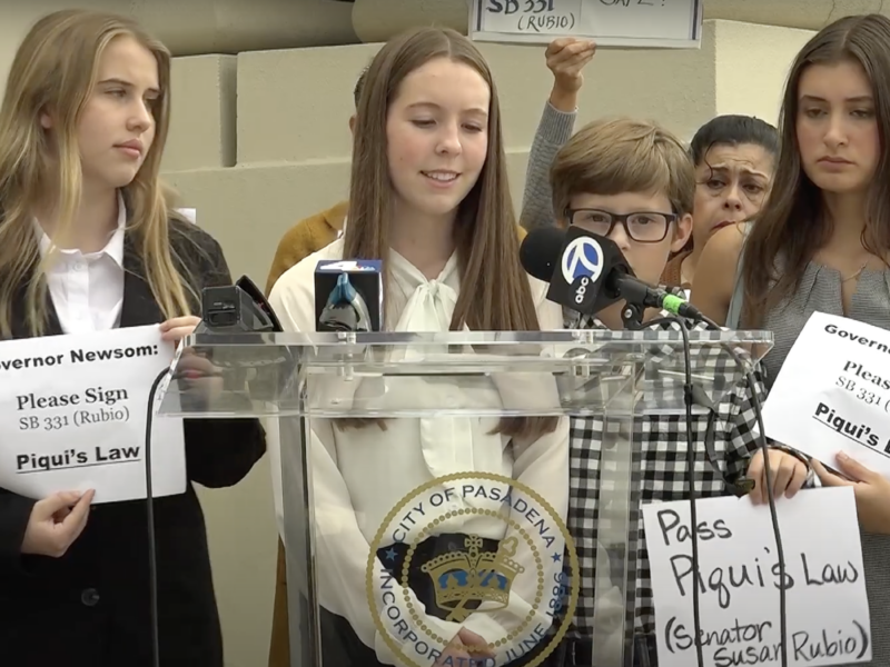 At a news conference about the passage of Piqui's Law, three teenage girls and a middle school-aged boy stand behind a clear lectern holding microphones from news outlets and bearing the seal of the city of Pasadena. They and others in the crowd hold signs, some of which read "Governor Newsom: Please sign SB 331 (Rubio) Piqui's Law" and "Pass Piqui's Law (Senator Susan Rubio)." They are referring to a bill that was introduced by California State Sen. Susan Rubio.