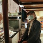 A man in his eighties sits on his bed in a small single-room occupancy residence in Chinatown. Behind him is a window where he hangs his clothes. There are assorted items beside him, including a desk fan, a television, newspapers and cardboard boxes.