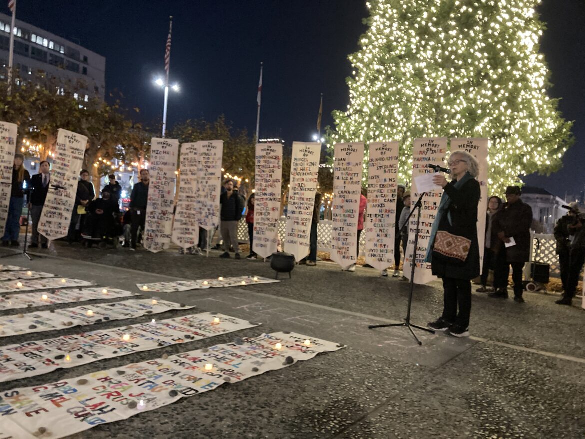 Several people stand in front of an evergreen tree covered in small white lights. They are holding vertical white banners displaying lists of people's names in colorful letters. Other banners are spread on the ground in front of them. A woman with shoulder-length gray hair wearing a black coat addresses the crowd speaking into a microphone on a stand.