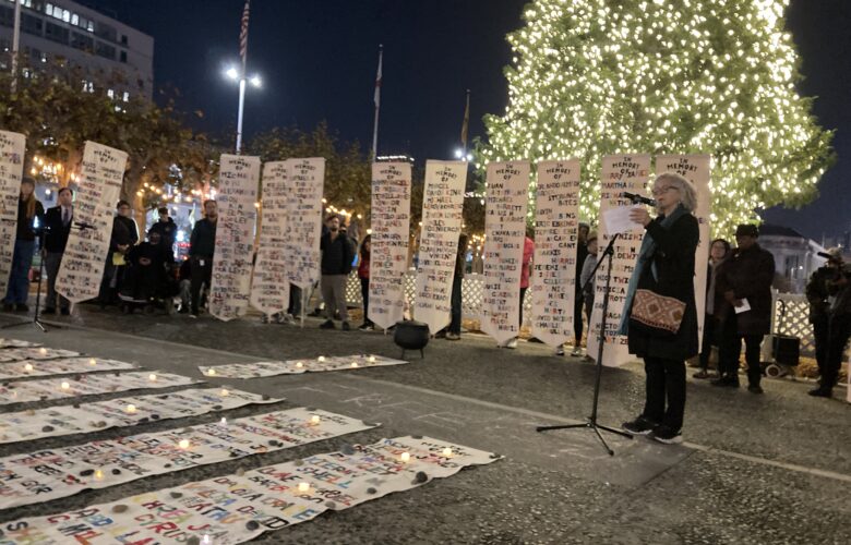 Several people stand in front of an evergreen tree covered in small white lights. They are holding vertical white banners displaying lists of people's names in colorful letters. Other banners are spread on the ground in front of them. A woman with shoulder-length gray hair wearing a black coat addresses the crowd speaking into a microphone on a stand.