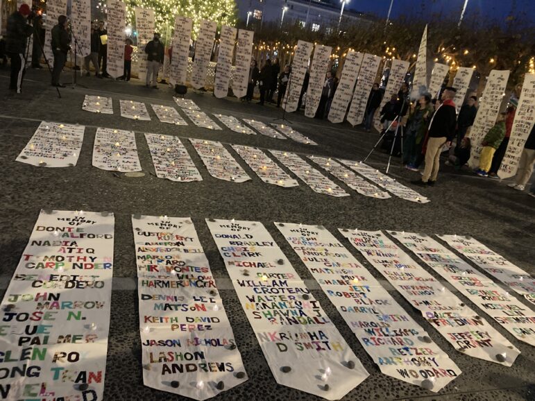 Long white banners covered in names in colorful lettering are displayed on the ground at a vigil for people who died this year in San Francisco while experiencing homelessness. Some banners are held up by people attending the event.