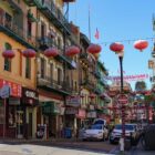 Red lanterns and flags are strung across the roadway on a block in San Francisco's Chinatown. Most of the three and four-story buildings have shops on the ground floor and apartments or offices above. Many of them have wrought iron balconies that are painted green.