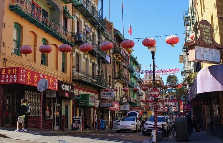 Red lanterns and flags are strung across the roadway on a block in San Francisco's Chinatown. Most of the three and four-story buildings have shops on the ground floor and apartments or offices above. Many of them have wrought iron balconies that are painted green.