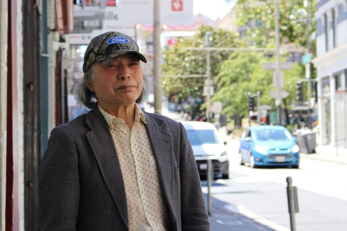 An older Asian man wearing a light colored button down shirt, a gray tweed sports coat and a baseball hat with the Ford automotive company logo on it stands facing the camera on an urban street with retail storefronts.