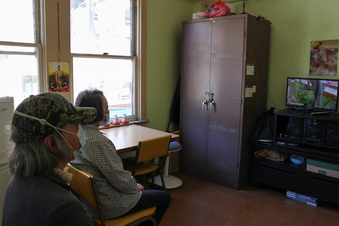 A man and a woman, both wearing surgical face masks, sit in a communal room watching a small flat-screen television. The walls are painted lime green.