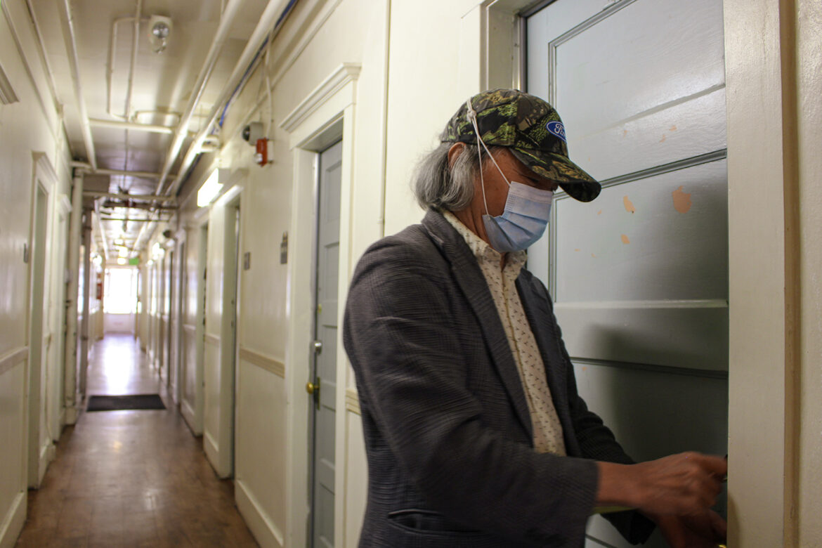 A man with gray hair wearing a light colored button-down shirt, a gray tweed jacket and a surgical mask uses a key to unlock a wooden door painted a light gray-blue in a long hallway filled with similar doors.
