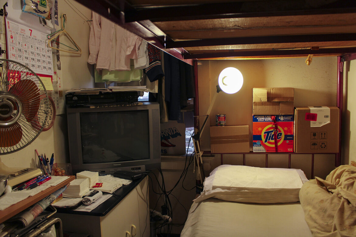 In a small crowded space, there is a bed, a table, fan, television and small low cabinet. Clothes hanging on hangers and personal belongings are stowed nearby.