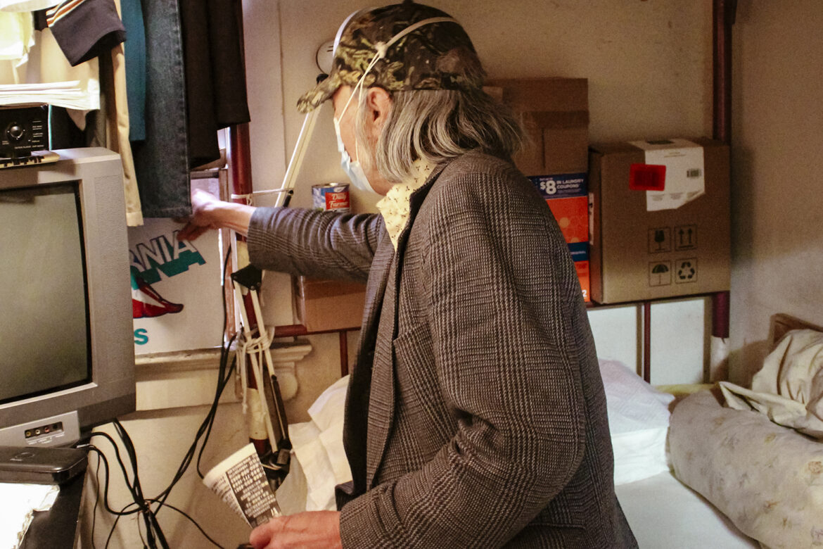 A man in a gray tweed jacket wearing a baseball cap and a surgical mask leans to adjust a piece of cardboard propped near the window in a small crowded room.