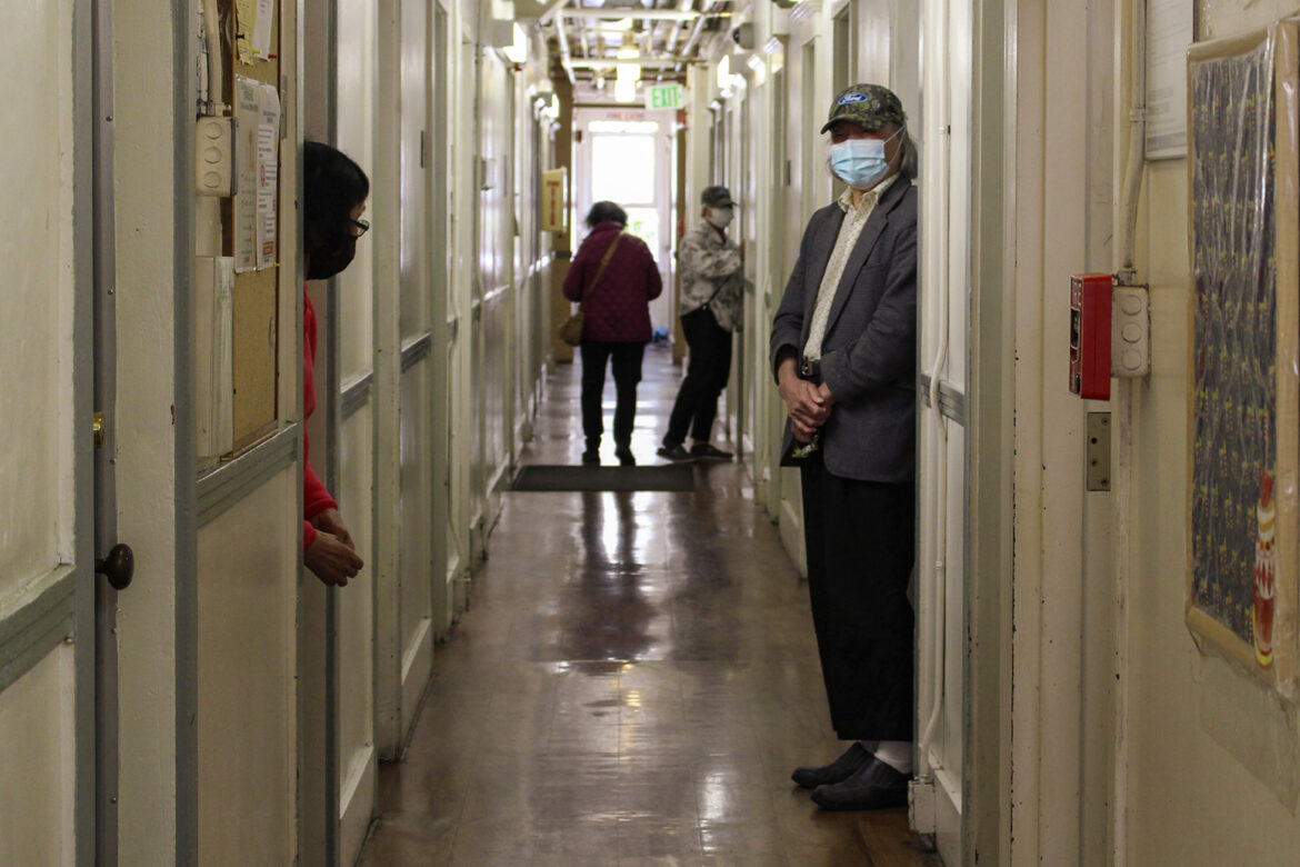 Two people stand in doorways, and two are walking at the far end of a long corridor filled with doorways. There is a window at the far end.