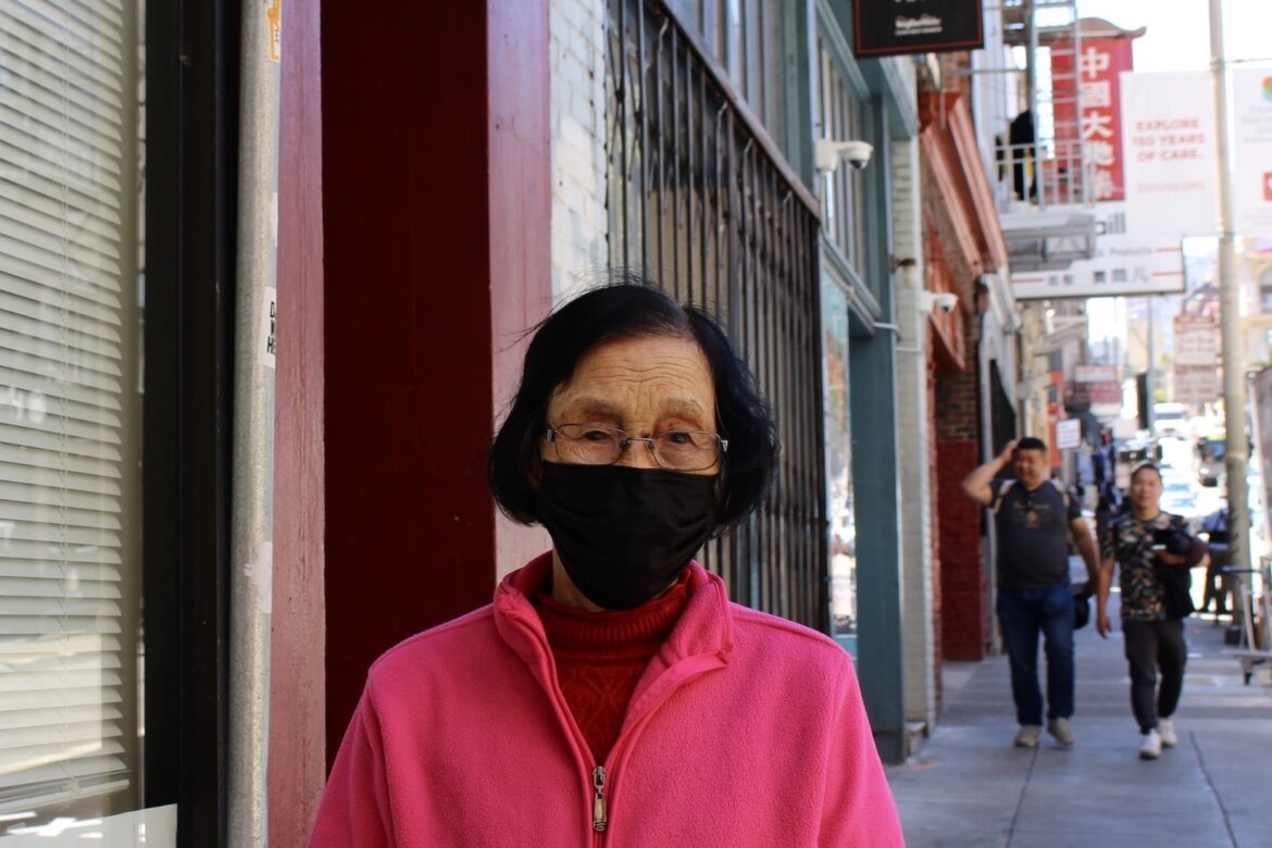 An older Asian woman with short dark hair wears a bright pink zip-up fleece jacket and a black face mask. She stands on the sidewalk of an urban street.