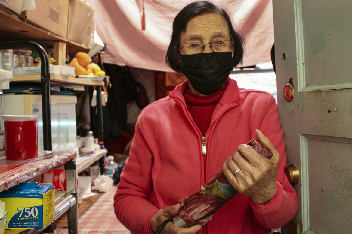 An older Asaisn woman with short dark hair wears a bright pink zip-up fleece jacket, glasses and a black face mask. She is standing in the entrance of a small, crowded room holding a small travel umbrella, rolled up and sheathed in a colorful sleeve