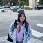 A young Latina with straight, long dark hair wears a lilac top and a jean jacket. She is smiling standing on a sidewalk near the intersection of a city street.