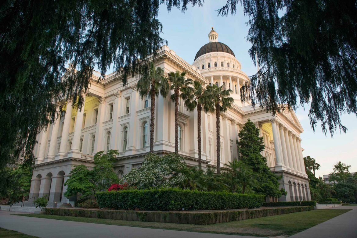 Exterior image of the California State Capitol building in Sacramento. Taken from a corner of the building at a low angle, the cream colored facade features columned entrances on each side, with the building's stately dome appearing toward the top of the frame. The building is surrounded by lush green landscaping.