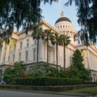 Exterior image of the California State Capitol building in Sacramento. Taken from a corner of the building at a low angle, the cream colored facade features columned entrances on each side, with the building's stately dome appearing toward the top of the frame. The building is surrounded by lush green landscaping.