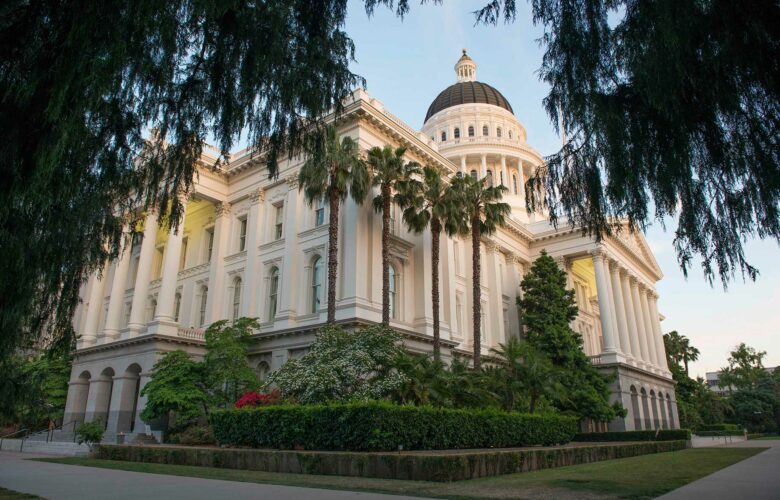 Exterior image of the California State Capitol building in Sacramento. Taken from a corner of the building at a low angle, the cream colored facade features columned entrances on each side, with the building's stately dome appearing toward the top of the frame. The building is surrounded by lush green landscaping.