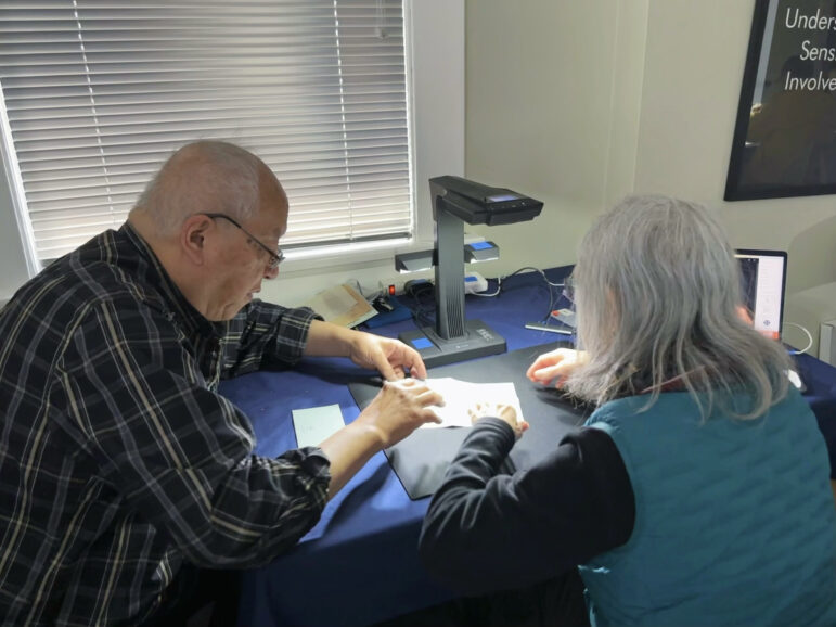 A man with short gray hair is in profile to the camera sitting next to a woman with shoulder-length gray hair sitting with her back to the camera. They are working together to place a document under the overhead scanner on the table in front of them.