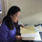A woman with dark shoulder length hair, and wearing a purple pullover and glasses, sits at a small table reviewing a small stack of documents.