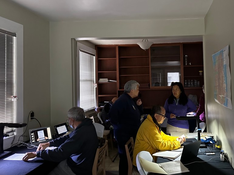 About half a dozen men and women gather in a dimly lit room with several tables. The back wall is lined with wooden bookshelves. In the foreground, two men sit at tables scanning documents.