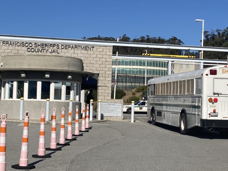 A white bus is stopped near the entrance of the San Francisco County Jail building in San Bruno. A sheriff's deputy stands in the shade near a one-story gatehouse with rounded concrete walls that is situated in front of a taller wall of sandstone-colored blocks. A row of tall orange and white traffic cones block access to another paved area in the foreground.