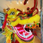 A large decorative yellow parade dragon head with multicolored accents appears with an open mouth featuring a pink and red paper mache tongue and large white teeth with fangs.